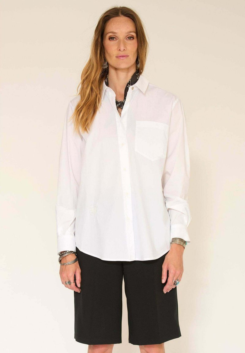 Chemise blanche à boutonnière avec une poche à l'avant, associée à un short noir. La chemise présente un col classique et des manches longues.