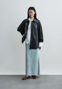 Camisa oversized preta em couro com dois bolsos no peito, combinada com uma saia de ganga azul claro e sapatos planos bordô, sobre um fundo neutro.