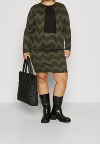Set šatů a svetru se zeleným vlnkovým vzorem; černý top a černé kozačky nad kolena; drží černou strukturovanou tašku na rameno.
