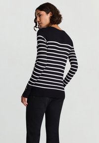 Schwarz-weiß gestreifter Pullover mit langen Ärmeln, rundem Ausschnitt und Knopfdetails an den Manschetten, kombiniert mit einer schwarzen Hose.