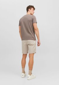 Camiseta de hombre ajustada marrón combinada con pantalones cortos beige. La camiseta tiene un cuello redondo; los pantalones cortos están doblados en el dobladillo. Se lleva con calcetines beige y zapatillas blancas.