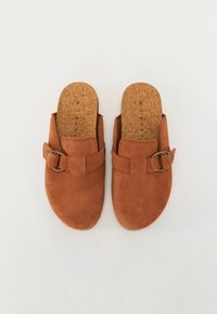 Brown suede slip-on mules with a cork insole, featuring a buckled strap accent on the upper. Flat sole design for comfort.