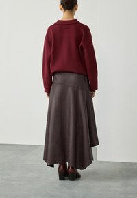 Donna che indossa un maglione bordeaux, una gonna in pelle asimmetrica marrone scuro e stivali con tacco abbinati, in piedi su un pavimento grigio contro un muro bianco.