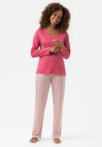 Femme souriante aux cheveux bouclés, pieds nus, portant un haut à manches longues rose et un pantalon de pyjama floral rose pâle, se tenant devant un fond blanc.