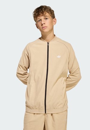 Chaqueta de terciopelo beige con cremallera negra, bolsillos laterales y pequeño logo blanco en el lado izquierdo del pecho. Textura suave, corte casual.