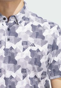 Poloshirt mit kurzen Ärmeln in einem weißen und marineblauen geometrischen Muster, mit einem Knopfkragen und einem Adidas-Logo. Hergestellt aus leichtem Stoff.