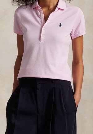 Persoon draagt een lichtroze poloshirt met korte mouwen en een klein geborduurd logo, gecombineerd met zwarte plissébroek met hoge taille.
