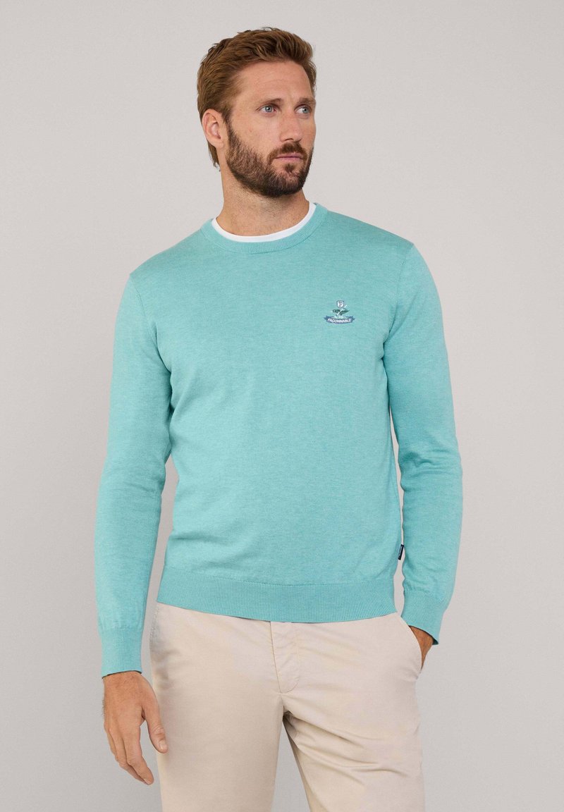 Jasnoniebieski dziany sweter z okrągłym dekoltem i ściągaczami. Posiada małe haftowane logo na piersi. Noszony z jasnymi spodniami.