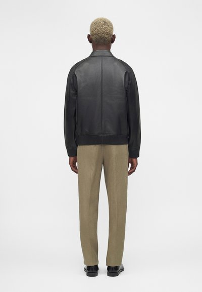 Άνδρας με κοντά ξανθά μαλλιά, που φοράει μαύρο δερμάτινο μπουφάν, μπεζ παντελόνια και μαύρα παπούτσια, στέκεται με την πλάτη γυρισμένη σε λευκό φόντο.