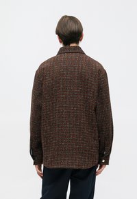 Brun og orange mønstret jakke med krave, lange ærmer og knapmanchetter. Struktureret stof med tydelige horisontale og vertikale linjer.