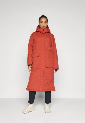 Woman standing wearing a long rust-orange hooded winter coat, black pants, white socks, and beige sneakers against a plain white background.
