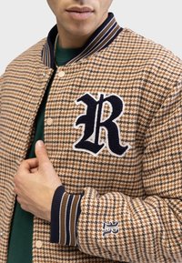 Veste de varsity à motif pied-de-poule marron avec des poignets et un col en maille bleu marine. Comprend un grand patch "R" bleu marine et des fermetures à pression en métal.