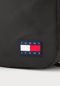 Svart ryggsekk med et rektangulært marineblått merke med teksten "TOMMY JEANS" og et rød og hvit logo-design. Glatt materiale, enkel design.