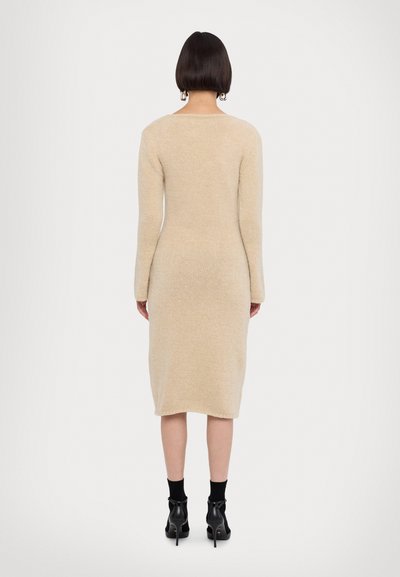 Abito a maglia beige con maniche lunghe, dalla silhouette aderente e lunghezza sotto al ginocchio. Texture liscia, collo a giro e dettaglio sul retro visibile. Stivali neri alla caviglia.