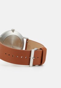 Brun läderarmband med en slät yta och spänne i silvermetal. Rund klockkapsel i rostfritt stål med en polerad yta.