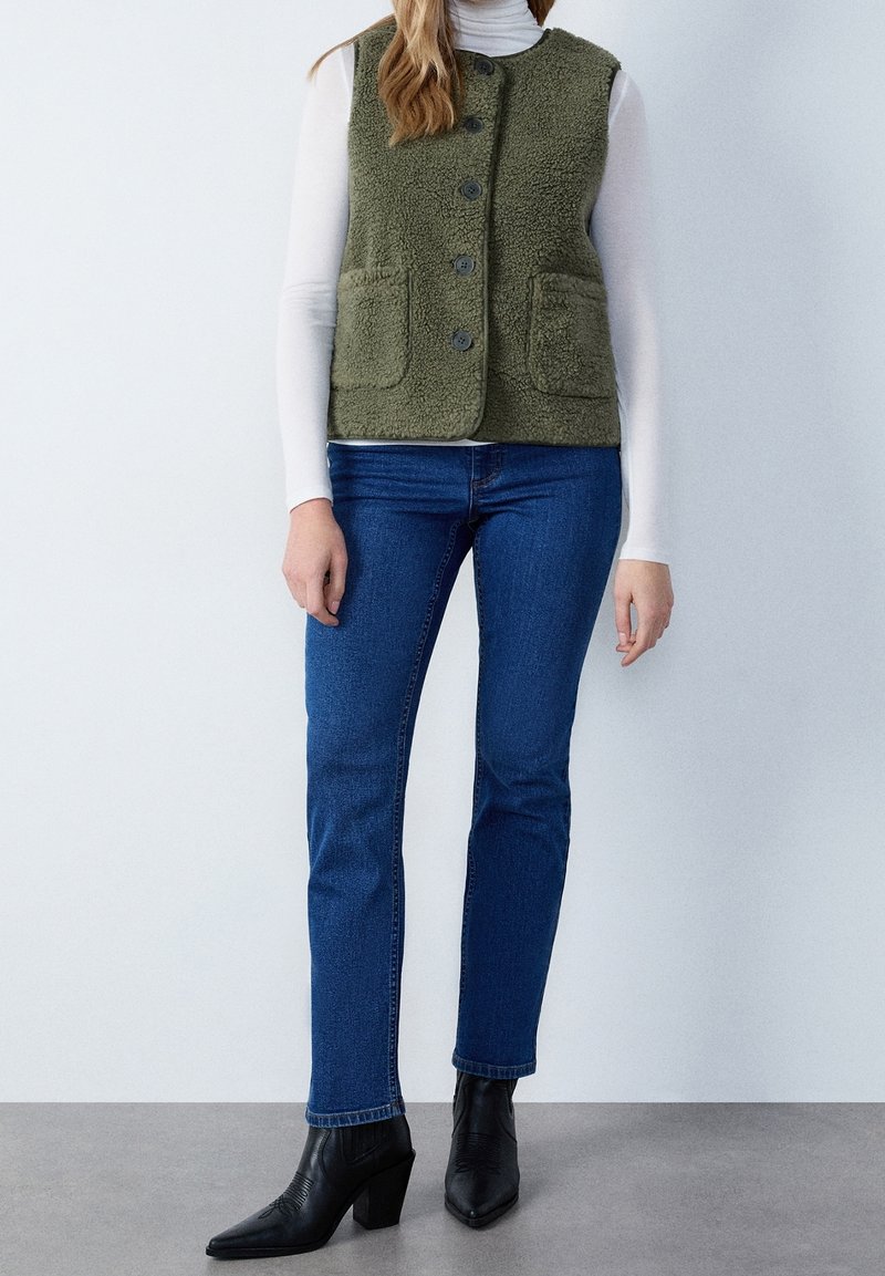 Frau, die eine grüne wattierte Weste mit Knöpfen, ein weißes langärmliges Rollkragenoberteil, blaue Jeans und schwarze Absatz-Stiefeletten trägt, vor einem einfarbigen Hintergrund.