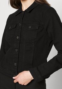 Musta farkkutakki, jossa on perinteinen kaulus, etupuolen läppätaskut, nappisuljenta ja pitkät hihat, joissa on kapea muoto.