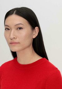 Maglione rosso con scollatura rotonda e rifiniture a coste, realizzato in un tessuto morbido. Il modello ha lunghi capelli neri lisci e un trucco minimale.
