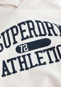 Světle zbarvená mikina s kapucí, na které je vysoce kontrastní námořnický text "SUPERDRY" a "ATHLETIC 72" spolu s grafickým symbolem skateboardu. Klasický design přes hlavu.