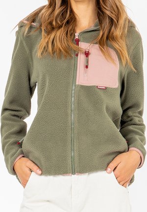 Persona che indossa una giacca in pile verde oliva con tasca con cerniera rosa e le mani nelle tasche dei pantaloni bianchi, mostrata dal collo ai fianchi.