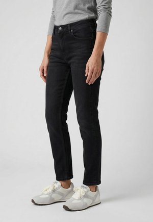 Osoba w czarnych obcisłych jeansach, szarej bluzce z długim rękawem i białych trampkach stoi na tle jednolitego jasnego tła.