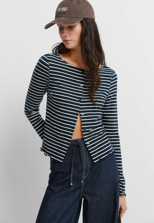 Donna che indossa un berretto marrone, una camicia a righe bianche e blu navy con maniche lunghe e bottoni, e jeans a vita alta di colore blu scuro.