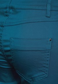 Tyrkysové látkové šortky s zadním záplátovým kapsou, plochými švy a kovovým knoflíkovým detailem. Hladká textura a přiléhavý střih.