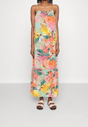 Woman wearing a sleeveless, colorful floral maxi dress with spaghetti straps and white crisscross sandals, standing against a plain background.