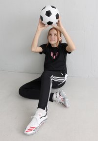 Dívka sedící na podlaze, držící nad hlavou černobílý fotbalový míč, oblečená v černém sportovním oblečení a bílých kopačkách, na pozadí bílé zdi.