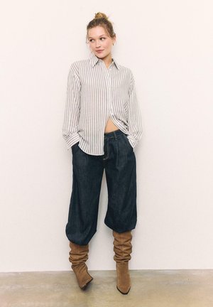 Jonge vrouw die een loszittend gestreept shirt draagt, donkere wijde jeans die in slordige tan suède laarzen zijn gestopt, staand tegen een effen muur.