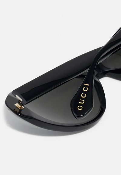 Sorte solbriller med vinkelret, kattetøjsform; mørke linser; blank overflade; guldfarvet "GUCCI" logo på armen; minimal detaljering.