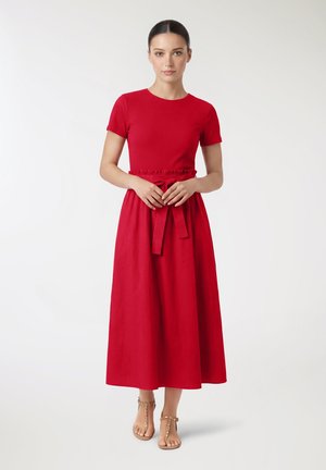 Donna in piedi che indossa un vestito rosso a maniche corte con cintura in vita e sandali beige con borchie, su sfondo bianco semplice.