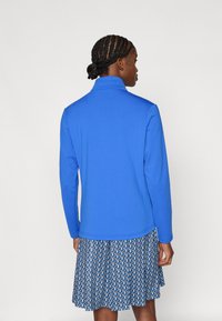 Zila flīsa jaka ar augstu apkakli, garām piedurknēm, teksturētu audumu un gludu apdari; uzvilkta pār rakstainu svārku.