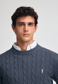 Mörkgrå kabelstickad tröja med ribbad halsringning, över en ljusblå skjorta med krage; har en liten broderad logotyp på bröstet.