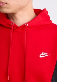 Prim-plan al unei persoane care poartă un hanorac roșu Nike cu șnururi vizibile și logo-ul alb Nike pe piept.