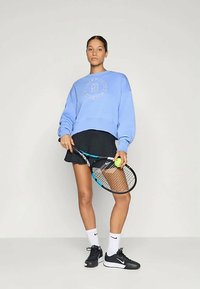 Γυναίκα με μπλε φούτερ και μαύρη τενιστική φούστα κρατάει ρακέτα και μπάλα τένις, στέκεται μπροστά από απλό φωτεινό φόντο.