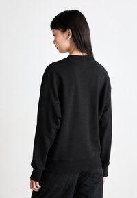 Črna oversize majica s dolgimi rokavi in rebrastimi manšetami, z okroglim izrezom in mehko teksturo tkanine. Zadnji pogled z minimalnimi detajli.