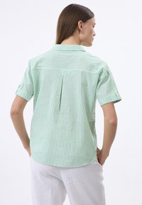 Camisa de manga corta a rayas verdes y blancas con dobladillo curvado, pliegue trasero y cuello con solapa. Fabricada en tejido ligero.