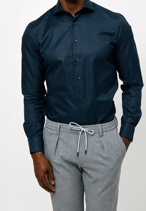 Homme portant une chemise bleu foncé ajustée boutonnée rentrée dans un pantalon gris clair à cordon, les mains détendues le long du corps.