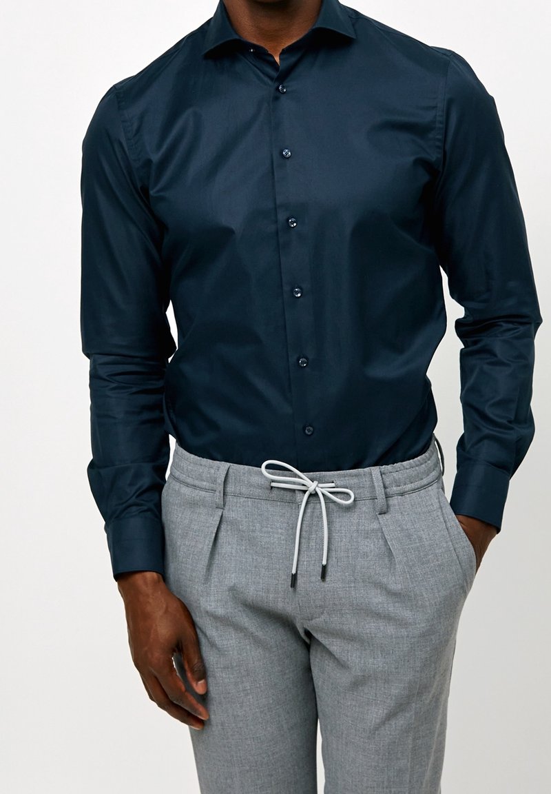 Homme portant une chemise bleu foncé ajustée boutonnée rentrée dans un pantalon gris clair à cordon, les mains détendues le long du corps.