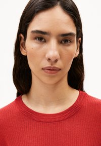 Rode geribbelde gebreide trui met een ronde halslijn, gemaakt van een textuurstof. Close-up die de stiksels en natuurlijke huidskleuren benadrukt.