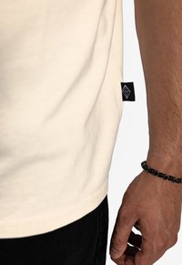 Beige bavlněné tričko s rovním střihem, na boku s černou visačkou. Na zápěstí je vidět černý korálkový náramek.