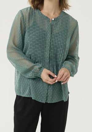 Femme portant un chemisier vert semi-transparent à manches longues avec des pois texturés, des poignets boutonnés et un devant plissé, associé à un pantalon noir.