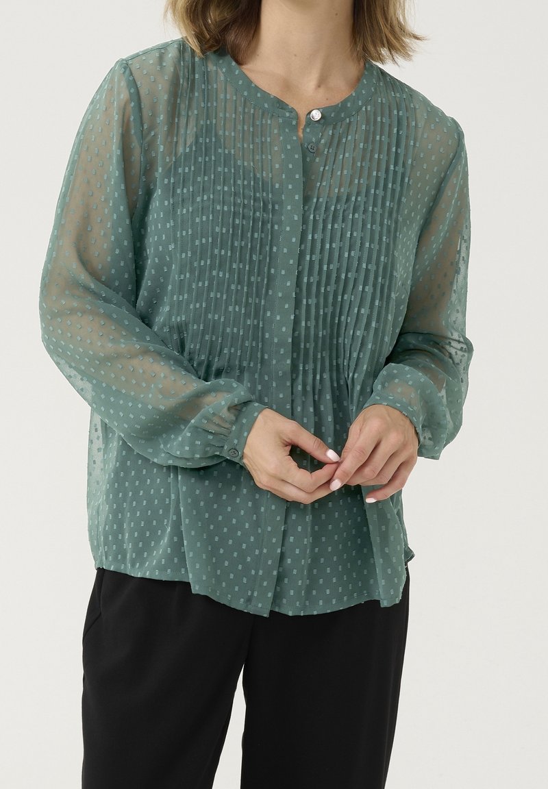 Femme portant un chemisier vert semi-transparent à manches longues avec des pois texturés, des poignets boutonnés et un devant plissé, associé à un pantalon noir.