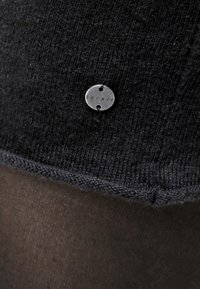 Tessuto a maglia nero con una superficie testurizzata visibile, caratterizzato da un'etichetta circolare d'argento con la scritta "ESPRIT" in rilievo. Accentuato da uno strato marrone liscio sottostante.