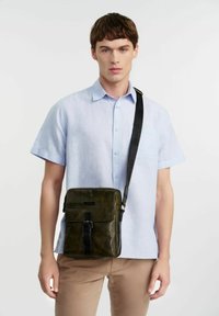 Zielona skórzana torebka na ramię o gładkiej fakturze, prostokątnym kształcie, z zapinanym na klapkę zapięciem oraz czarnym regulowanym paskiem. Z przodu znajduje się elegancka klamra.