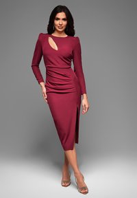 Robe bordeaux à manches longues avec une fente sur le côté et un col découpé. Tissu lisse, silhouette ajustée avec des détails froncés sur le côté.