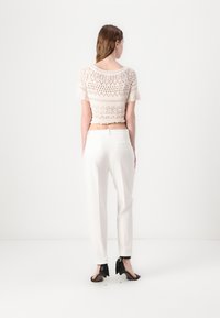 Háčkovaný crop top v béžovej farbe s krátkymi rukávmi a otvoreným vzorom; v kombinácii s bielymi strihanými nohavicami a čiernymi sandálmi na vysokom opätku so stužkami.
