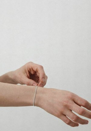 Persoană care închide o brățară delicată din lanț de argint la încheietura mâinii stângi, pe un fundal simplu, deschis la culoare.