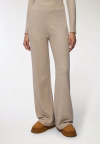 Beżowe, prążkowane spodnie dzianinowe z wysokim stanem i szerokimi nogawkami, wykonane z miękkiego, teksturowanego materiału. Stylizowane z brązowymi butami wsuwanymi.